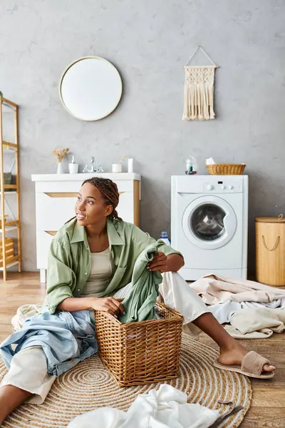Афроамериканка с афрокосичками сидит на полу в ванной комнате с корзиной для белья, делает домашнюю работу. — стоковое фото