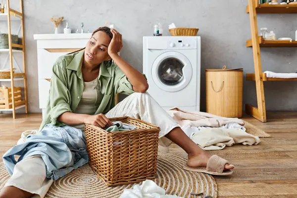 Una mujer afroamericana con trenzas afro se sienta al lado de una canasta de ropa en el baño, preparándose para lavar la ropa. - foto de stock