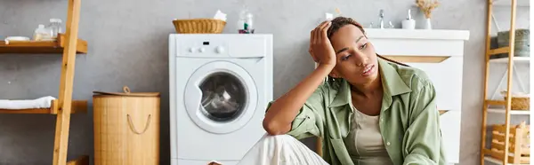 An African American woman with afro braids sits on the floor, with a laundry machine in the background, doing housework. — Stock Photo