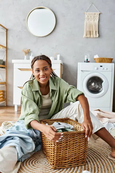 Una mujer afroamericana con trenzas afro está sentada en el suelo, rodeada por una cesta llena de ropa, lavando su ropa. - foto de stock