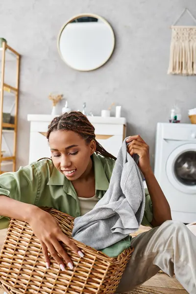 A woman of African American descent, with stylish afro braids, sits on a bed while gently holding a woven basket. — Stock Photo