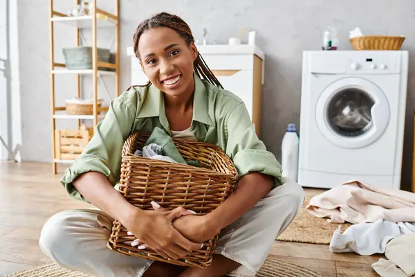 Uma mulher afro-americana com tranças afro ajoelha-se no chão, segurando graciosamente uma cesta enquanto lava a roupa no banheiro. — Fotografia de Stock