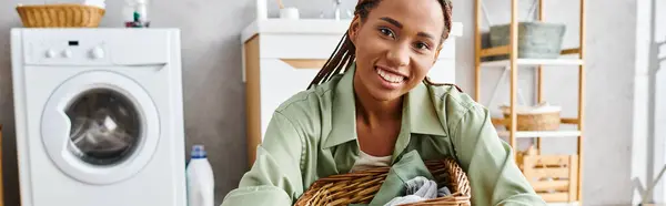 An African American woman with afro braids holding a basket full of laundry in a bathroom. — Stock Photo