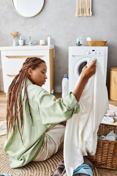 Une Afro-Américaine avec des tresses afro est assise sur le sol à côté d'une machine à laver, se concentrant sur la blanchisserie dans la salle de bain. — Photo de stock