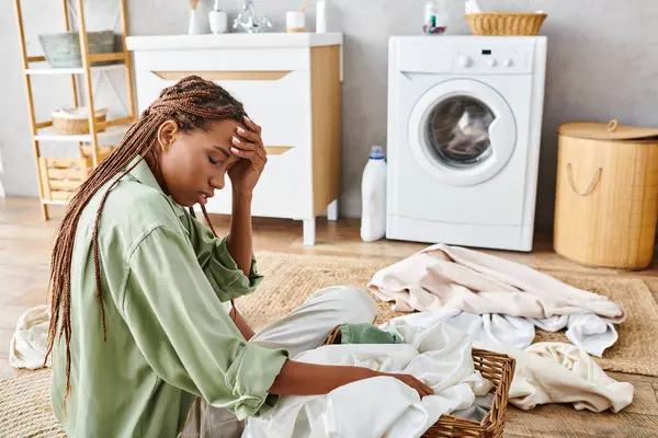 Une femme avec des tresses afro s'assoit à côté d'un tas de vêtements, perdu dans la pensée tout en faisant la lessive dans une salle de bain. — Photo de stock