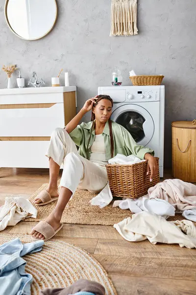 Una mujer afroamericana con trenzas afro se sienta junto a una canasta de ropa en un baño, dedicada a las tareas domésticas. - foto de stock