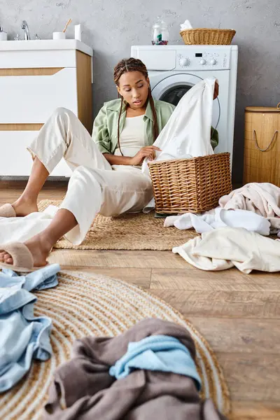 Una mujer afroamericana con trenzas afro se sienta en el suelo junto a una pila de ropa sucia, profundamente pensada en medio de la tarea del hogar. - foto de stock