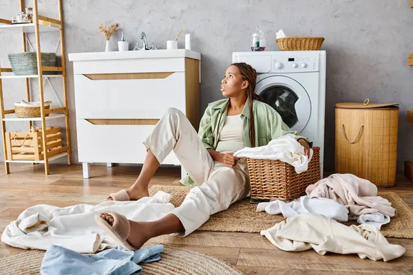 Una mujer afroamericana con trenzas afro sentada en el suelo frente a una lavadora, lavando ropa en el baño. - foto de stock