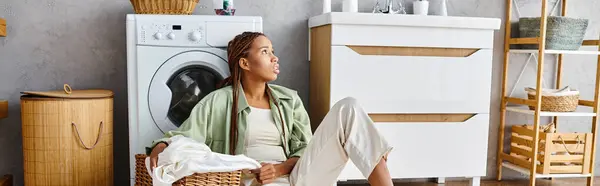 Mujer afroamericana con trenzas afro se sienta frente a una lavadora, lavando ropa en un baño. - foto de stock