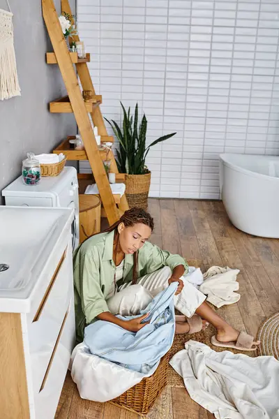 Una mujer afroamericana con trenzas afro se sienta en el piso del baño, rodeada de lavandería mientras hace las tareas domésticas. - foto de stock