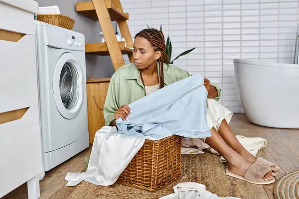 Una mujer afroamericana con trenzas afro se sienta en el suelo junto a una canasta de ropa en un baño, clasificando la ropa. - foto de stock