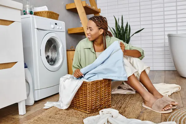 An African American woman with afro braids sits next to a washing machine, tending to laundry in a cozy bathroom setting. — Stock Photo