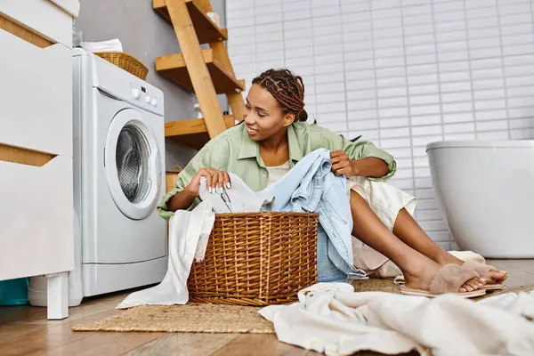 Femme afro-américaine avec des tresses afro assis à côté d'une machine à laver, faire la lessive dans un cadre de salle de bains. — Photo de stock