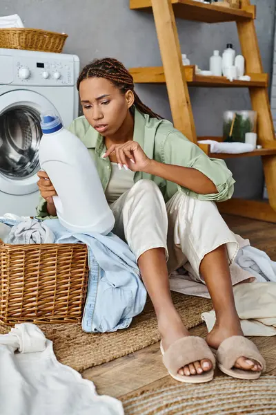 African American woman with afro braids doing laundry sits on floor next to a washing machine in a bathroom. — Stock Photo
