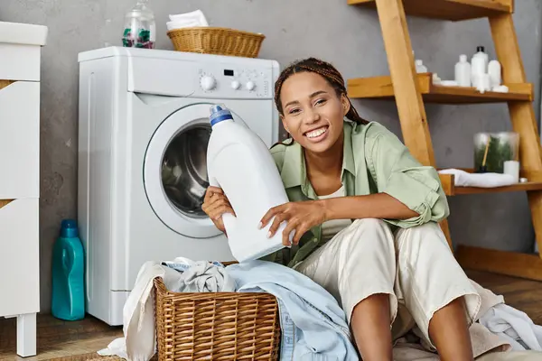 An African American woman with afro braids sits next to a washing machine, doing laundry in a bathroom setting. — Stock Photo