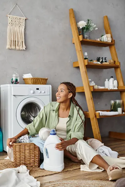 Una mujer afroamericana con trenzas afro se sienta tranquilamente en el suelo junto a una lavadora, lavando la ropa en un baño. - foto de stock