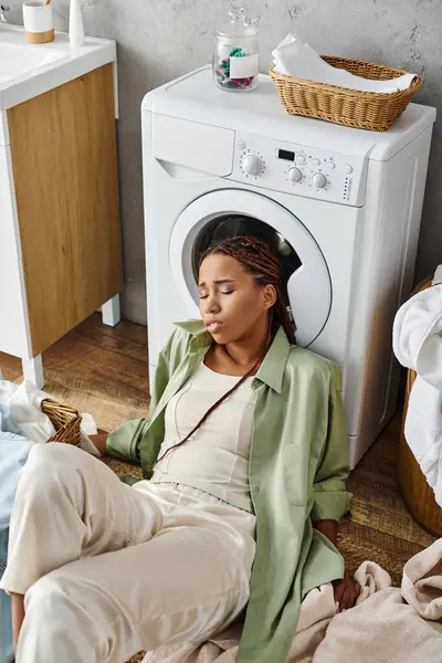 Africano mulher americana com tranças afro sentado ao lado de uma máquina de lavar roupa, fazendo lavanderia doméstica em um banheiro. — Fotografia de Stock