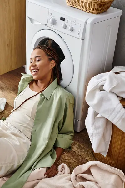 Uma mulher afro-americana com tranças afro senta-se no chão ao lado de uma máquina de lavar roupa, lavando a roupa no banheiro. — Fotografia de Stock