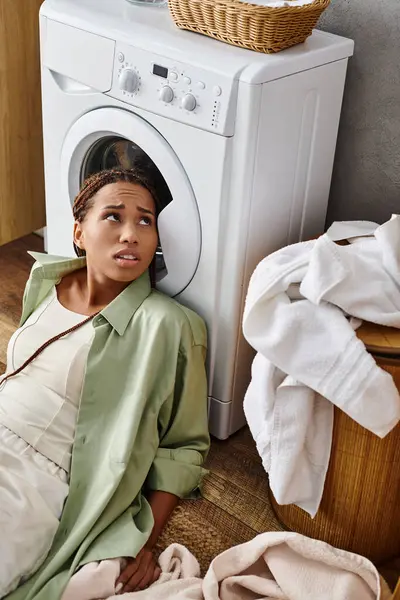 Una mujer afroamericana con trenzas afro sentada en el suelo junto a una lavadora, lavando la ropa en un baño. - foto de stock