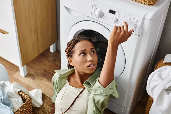 Una mujer afroamericana con trenzas afro mira hacia arriba a una secadora mientras hace la colada en un baño. - foto de stock