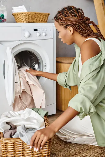 An African American woman with afro braids diligently loading clothes into a washing machine in a bathroom. — Stock Photo