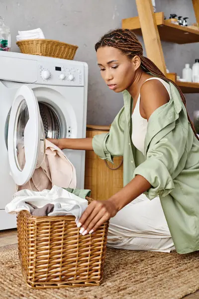 Femme afro-américaine avec des tresses afro trier et placer des vêtements dans un panier à côté d'une machine à laver dans une salle de bain. — Photo de stock