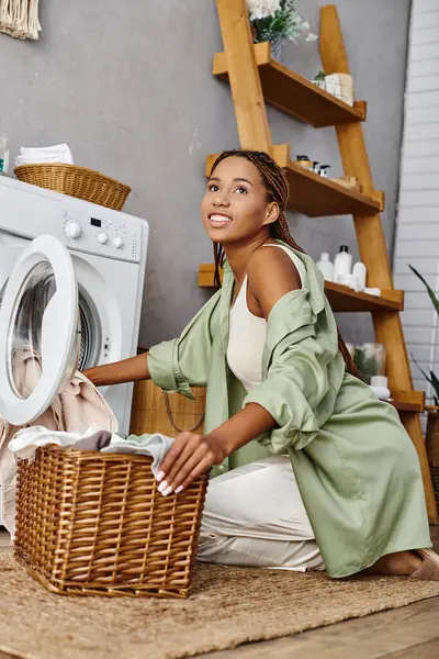 Une Afro-Américaine avec des tresses afro s'assoit sur le sol près d'une machine à laver, faisant la lessive dans un cadre de salle de bain. — Photo de stock