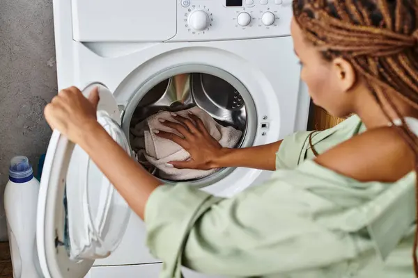 Uma mulher afro-americana com tranças energeticamente lavando roupa em um banheiro com uma máquina de lavar roupa. — Fotografia de Stock