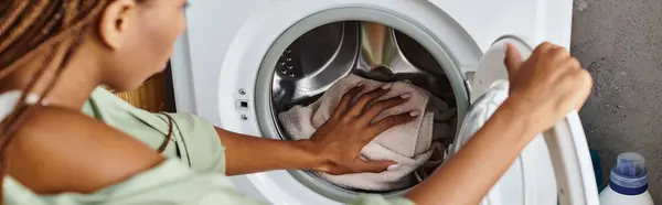 Una mujer afroamericana con trenzas afro limpia diligentemente la parte delantera de una lavadora en un baño. - foto de stock