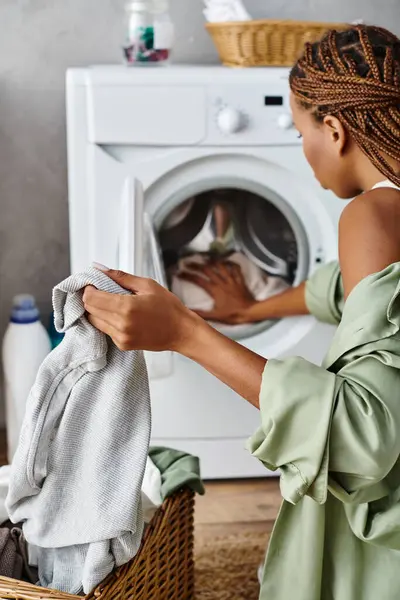 Eine afroamerikanische Frau mit Afro-Zöpfen legt in einem schön dekorierten Badezimmer fleißig Kleidung in einen modernen Trockner. — Stockfoto