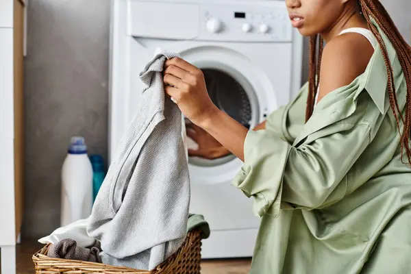 Una mujer afroamericana con trenzas afro se ve secando su ropa en una canasta de ropa en un baño. - foto de stock