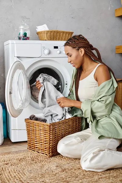 Una mujer afroamericana con trenzas afro sentada en el suelo junto a una lavadora, lavando la ropa en un baño. - foto de stock