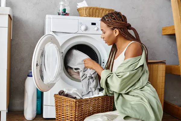Africano mulher americana com tranças afro sentado por uma máquina de lavar roupa fazendo lavanderia em um banheiro. — Fotografia de Stock