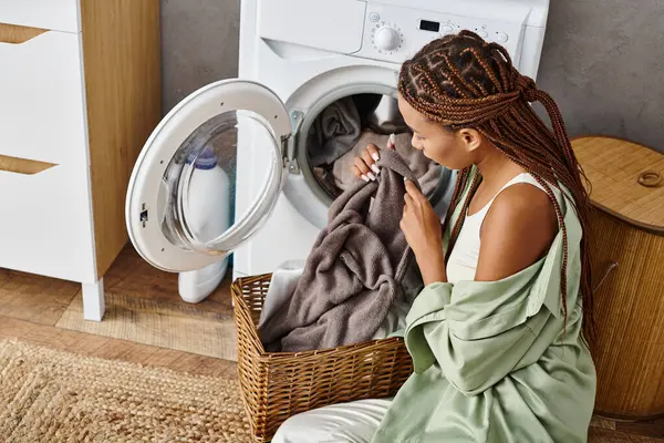 Eine afroamerikanische Frau mit Afro-Zöpfen sitzt vor einer Waschmaschine und wäscht fleißig im Badezimmer. — Stockfoto