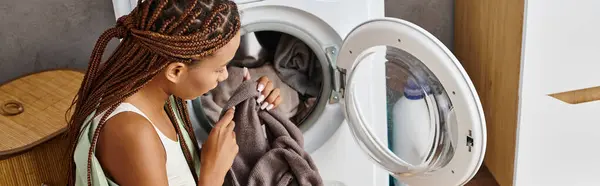 An African American woman with afro braids gazes at her reflection in a mirror while doing laundry in a bathroom. — Stock Photo