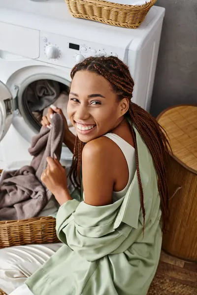Una mujer afroamericana con trenzas afro se sienta en el suelo junto a una lavadora, lavando la ropa en el baño. - foto de stock