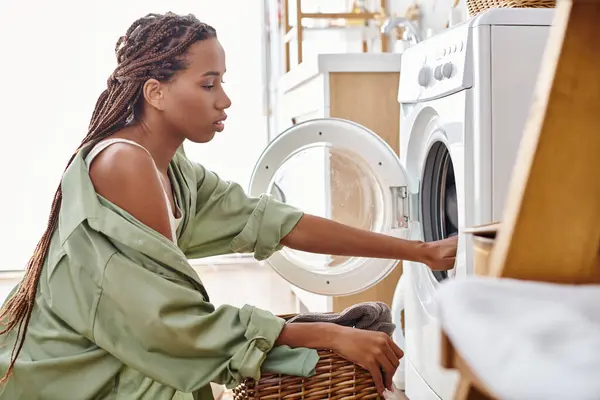 Una mujer afroamericana con trenzas afro carga una lavadora en una secadora mientras hace la colada en un baño. - foto de stock