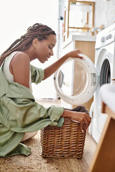 An African American woman with afro braids sits next to a washing machine in a bathroom, focused on doing the laundry. — Stock Photo