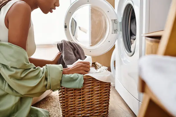 Афроамериканка с афрокосичками сортирует и кладет одежду в корзину рядом с стиральной машиной в ванной комнате. — стоковое фото