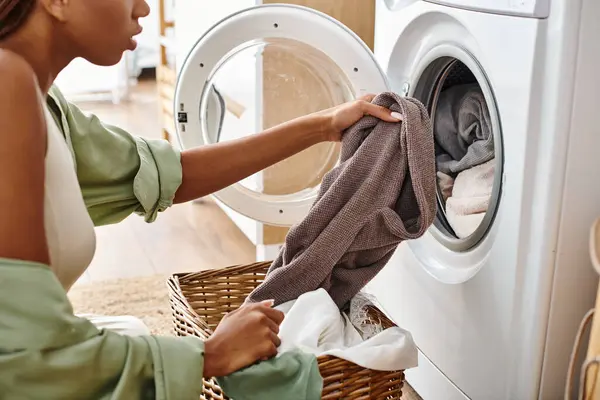 Donna afro-americana con trecce afro mettere un panno in un asciugatrice in un bagno durante il tempo di lavanderia. — Foto stock