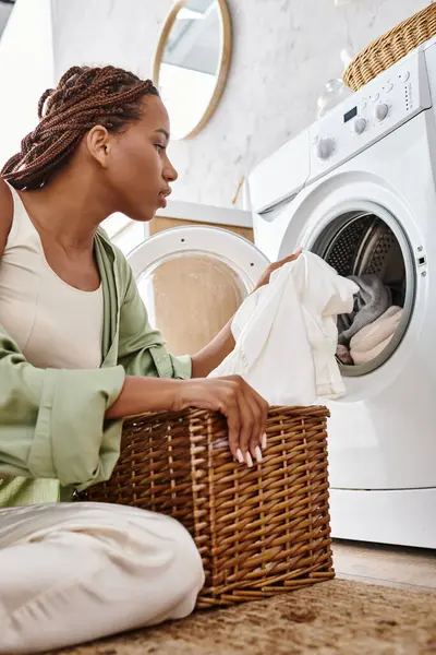 Uma mulher afro-americana com tranças se senta na frente de uma máquina de lavar roupa, lavando roupas em um banheiro. — Fotografia de Stock