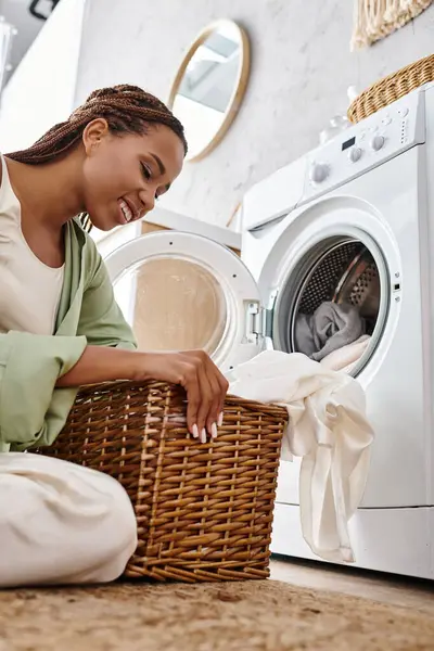 Femme afro-américaine avec des tresses afro assis sur le sol à côté de la machine à laver, faire la lessive dans la salle de bain. — Photo de stock