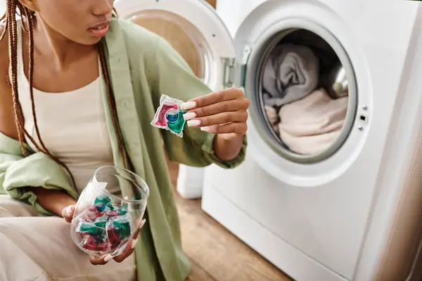 Uma afro-americana com tranças sentada ao lado de uma máquina de lavar roupa, lavando a roupa em um banheiro. — Fotografia de Stock