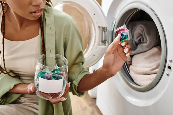 Uma mulher afro-americana com tranças afro desfruta de um momento tranquilo, segurando uma cápsula de gel junto a uma máquina de lavar roupa em um banheiro acolhedor. — Fotografia de Stock