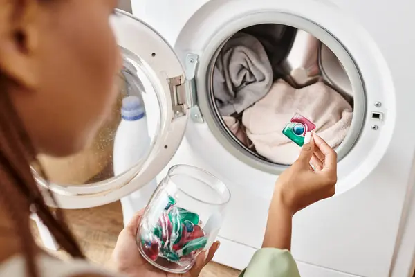 Une Afro-Américaine avec des tresses afro regarde la capsule de gel dans une machine à laver tout en faisant la lessive dans une salle de bain. — Photo de stock