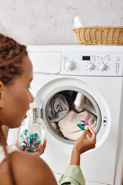 Une Afro-Américaine avec des tresses afro regarde à l'intérieur d'une machine à laver dans une salle de bain tout en faisant la lessive. — Photo de stock