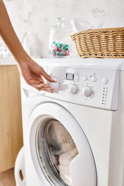 Femme afro-américaine ajustant un bouton sur une machine à laver tout en faisant la lessive dans une salle de bain. — Photo de stock