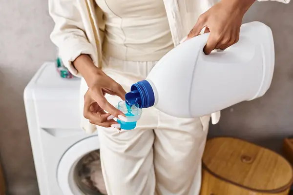 Femme afro-américaine avec des tresses afro tenant une bouteille de détergent tout en faisant la lessive près d'une machine à laver dans une salle de bain. — Photo de stock
