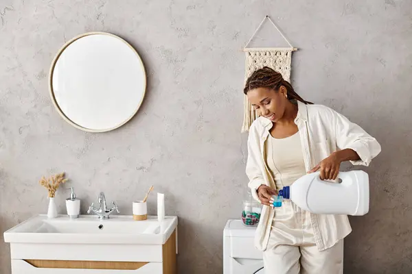 Una mujer afroamericana con trenzas afro está vertiendo pacíficamente detergente en un recipiente mientras lava la ropa en un baño. - foto de stock