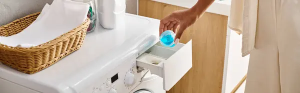 Uma mulher afro-americana limpa uma máquina de lavar roupa usando uma cápsula de gel azul em um banheiro. — Fotografia de Stock